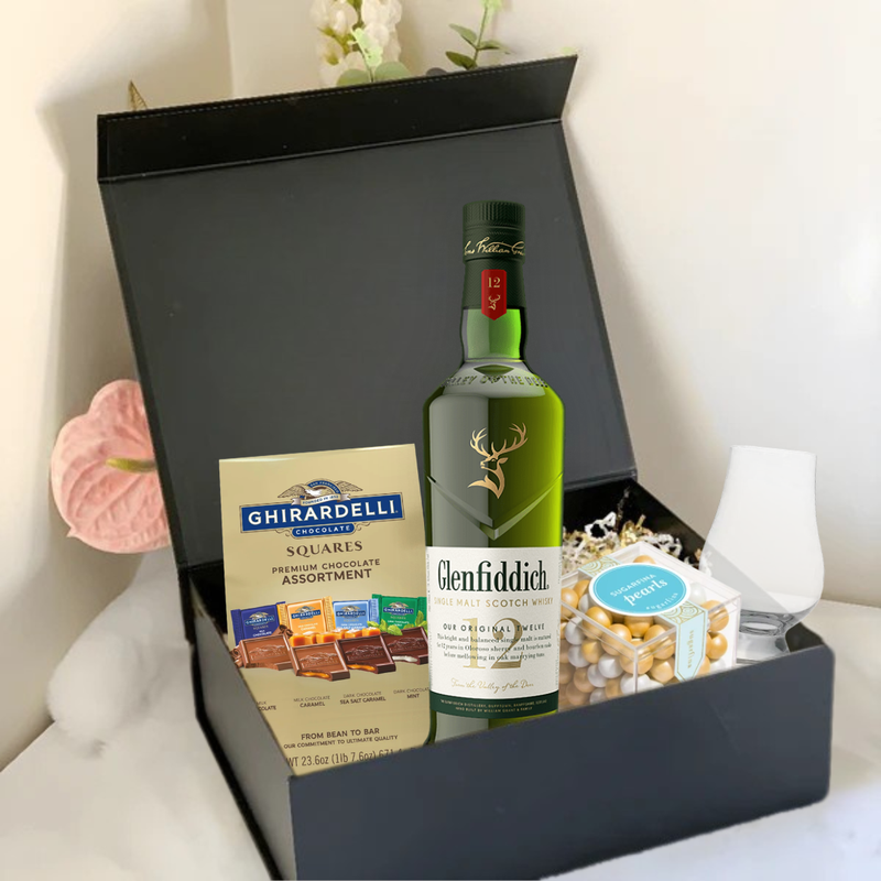 Load image into Gallery viewer, The Premium Scotch Gift Box by Main Street Liquor features Glenfiddich 12 Year Old whisky, Ghirardelli chocolate squares, a glass, and mixed nuts, all elegantly arranged with decorative flowers in the background.
