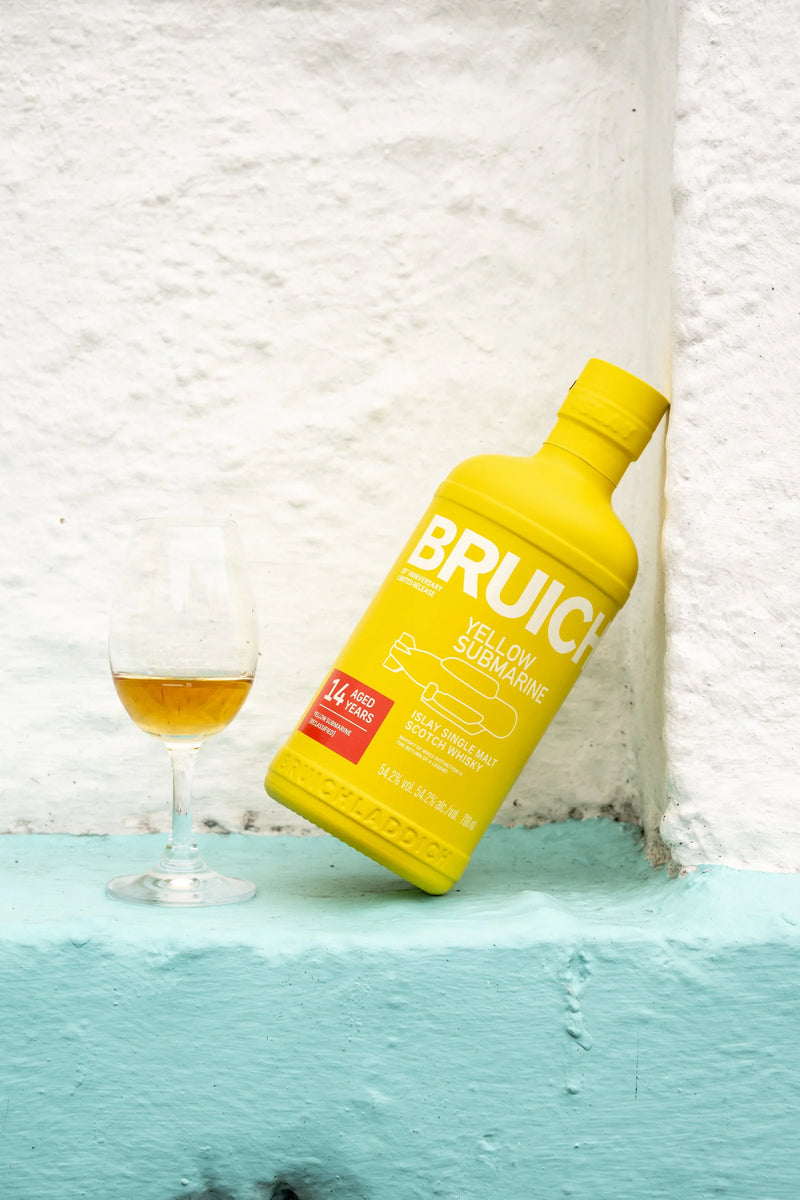 Load image into Gallery viewer, A yellow bottle of Bruichladdich Yellow Submarine III 14 Year Old Islay Single Malt Scotch Whisky leans against a white wall, beside a glass of amber liquid on a light blue ledge.
