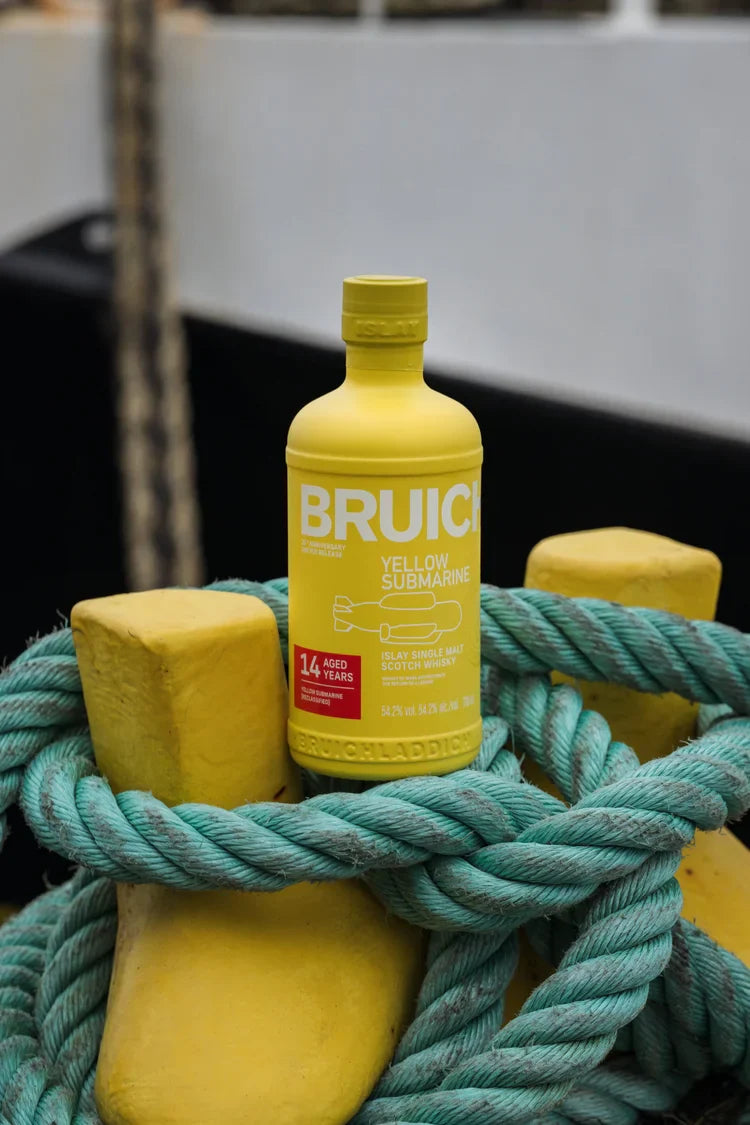 Load image into Gallery viewer, A yellow bottle of Bruichladdich Yellow Submarine III 14 Year Old Islay Single Malt Scotch Whisky sits on yellow bollards with teal rope, with an out-of-focus nautical background.
