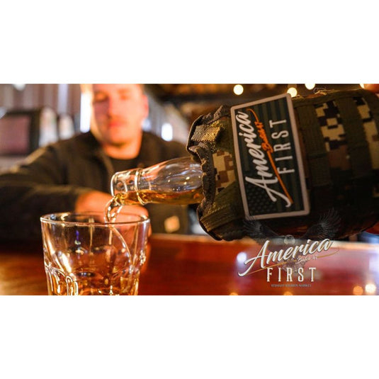 A close-up of a bottle in a camouflage sleeve labeled America First Straight Bourbon Whiskey pours rich bourbon into a glass, with a blurred figure at the bar—highlighting America First’s dedication to true American craftsmanship.