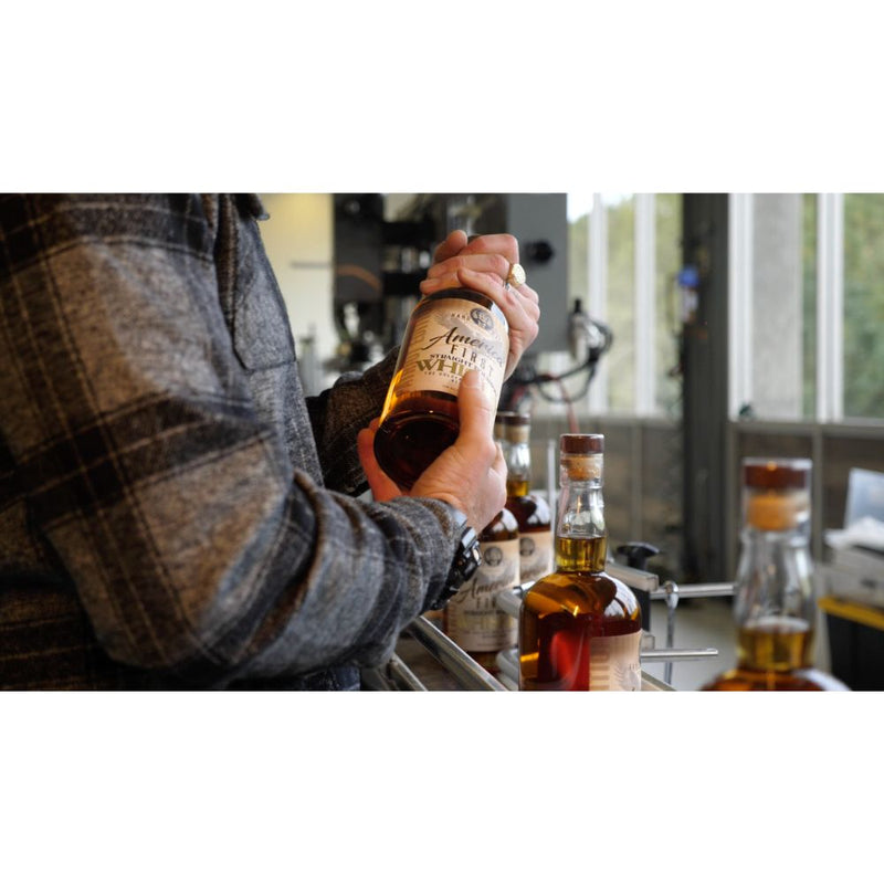Load image into Gallery viewer, Wearing a plaid jacket, a person holds an America First Straight Bourbon Whiskey 2 Pack at a bar, with other America First bottles highlighting American craftsmanship on the counter by large windows.
