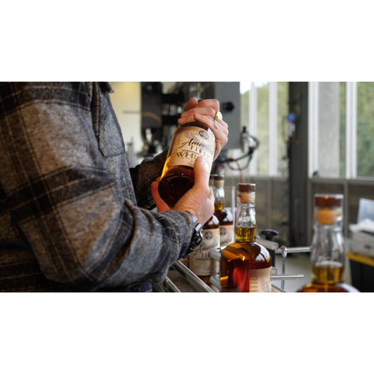 A person in a plaid jacket holds a bottle of America First Straight Bourbon Whiskey at a counter, surrounded by other whiskey bottles and large windows—an inviting showcase of American craftsmanship.