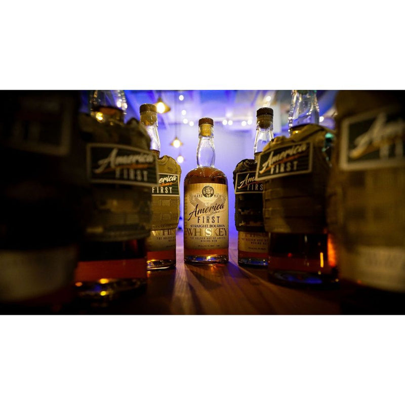 Load image into Gallery viewer, An America First Straight Bourbon Whiskey 6 Pack sits on a table, surrounded by other whiskey bottles, with warm string lights in the background creating a cozy, inviting atmosphere.
