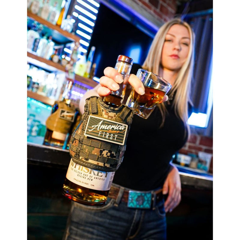 Load image into Gallery viewer, A woman at a bar holds a glass of whiskey and an America First Straight Bourbon Whiskey 3 Pack, the limited-edition set wrapped in camouflage. Bar shelves lined with bottles are visible in the background.

