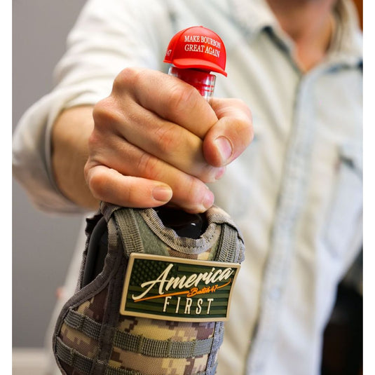 A person in a light-colored, long-sleeved shirt holds an America First Straight Bourbon Whiskey 6 Pack bottle with a red cap reading MAKE BOURBON GREAT AGAIN, featuring a limited-edition camouflage sleeve and America First patch.