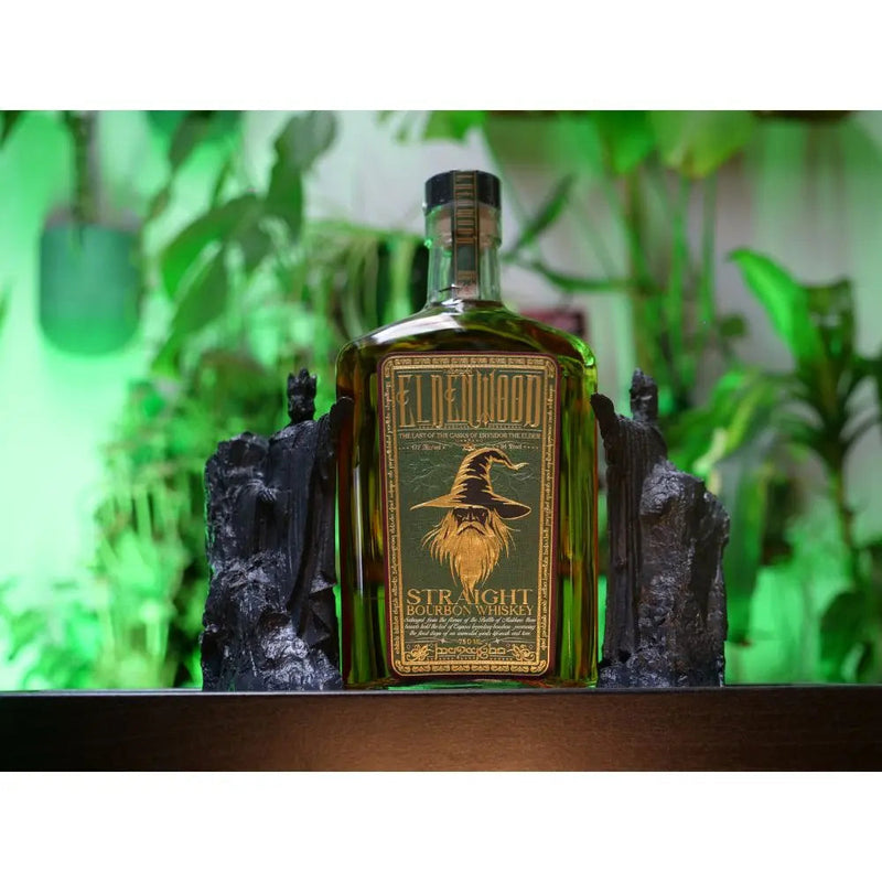Load image into Gallery viewer, A bottle of Eldenwood Straight Bourbon Whiskey by Eldenwood, with Eryndor the Elder on the label, is placed between two dark bookends and framed by lush green plants in the background.
