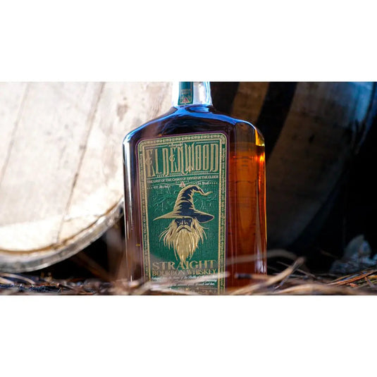 A bottle of Eldenwood Straight Bourbon Whiskey, with a green and gold label featuring Eryndor the Elder, a wizard illustration, set before a wooden barrel and surrounded by straw.