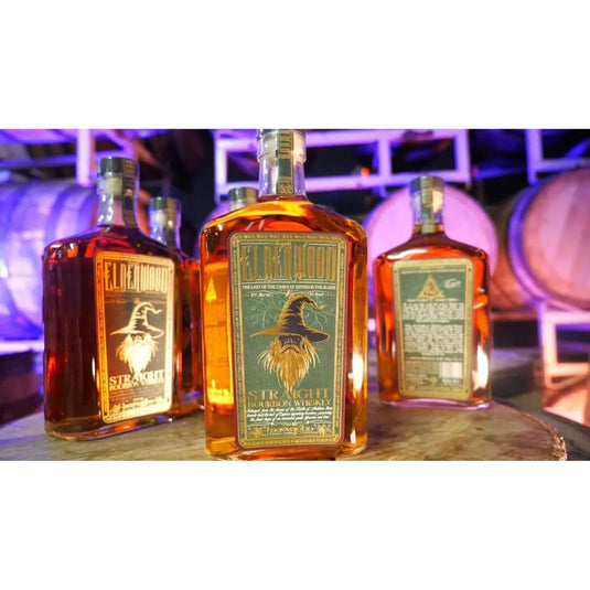 Three bottles of Eldenwood Straight Bourbon Whiskey 3 Pack sit on a barrel, with barrels behind and purple lighting. Each bottle from Eldenwood features a bearded wizard illustration on the label.