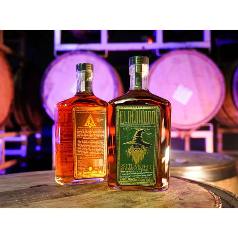 Load image into Gallery viewer, Two bottles of Eldenwood Straight Bourbon Whiskey sit atop a wooden barrel, with large oak barrels blurred in the background and purple lighting casting a glow over the scene.
