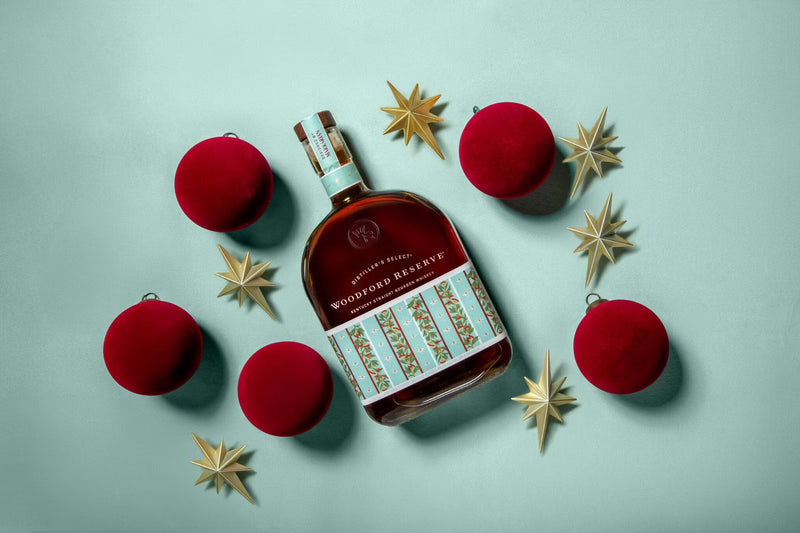 Load image into Gallery viewer, A Woodford Reserve Holiday Edition Bourbon 2025 “Designed by Markarian” 3 Pack is displayed amid red ornaments and gold star decorations, all set on a light blue background.
