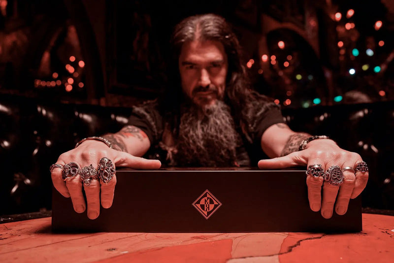Load image into Gallery viewer, A man with long hair and a beard sits at a table, gripping a Shotgun Blast Whiskey First Edition Straight Bourbon collector’s box. He wears ornate rings as dramatic reddish lighting casts a mysterious vibe, highlighting the unique Machine Head branded product.

