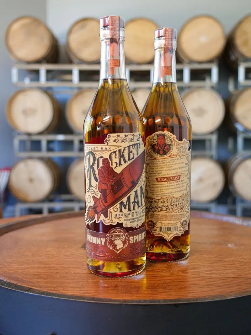 Load image into Gallery viewer, Two bottles of Rocket Man Bourbon Whiskey by Rocket Man, featuring limited release illustrated labels, rest on a barrel with rows of barrels in the background—an ideal scene for this political parody whiskey.
