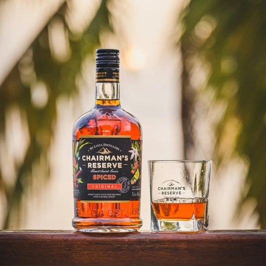 A bottle of Chairman's Reserve Spiced Original from Chairman's Reserve Rum and a glass of Caribbean rum rest on a wooden surface, framed by blurred palm leaves and sunlight in the background.