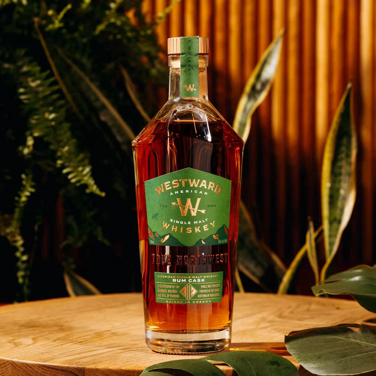 Load image into Gallery viewer, A bottle of Westward American Single Malt Rum Cask by Westward rests on a wooden surface, surrounded by green plants and warm wood tones. The label features green with gold and white text.
